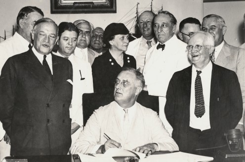 President Roosevelt signing the Social Security Act. Among those with the President are Senator Alben Barkley, Senator Robert Wagner, Senator Robert LaFollette, Secretary of Labor Frances Perkins, Representative Pat Harrison, and Representative David