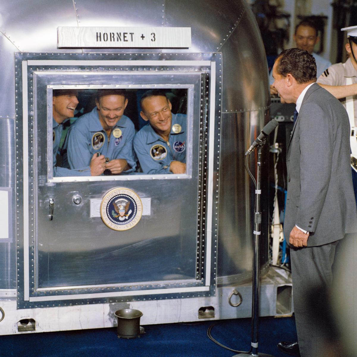 President Nixon speaks to the Apollo 11 astronauts aboard the USS Hornet.  The astronauts are in isolation.  The Hornet +3 is a reference to the 3 astronauts added to the USS Hornet's passengers and crew.