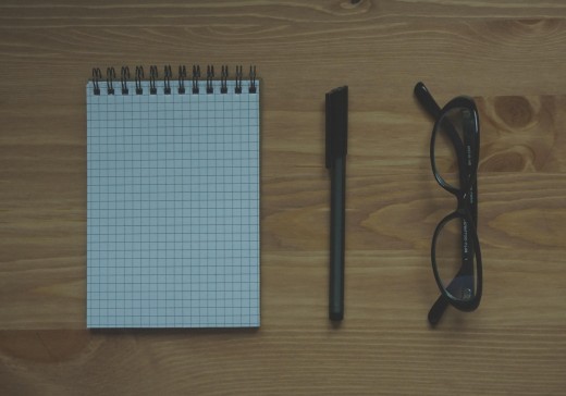 A notepad, pen, and glasses sit on a wooden table, shot from above.