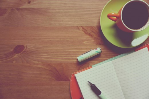 Wooden table, shot from above, with an orange mug on a green saucer, a notebook, and a pen and lid resting to the right.
