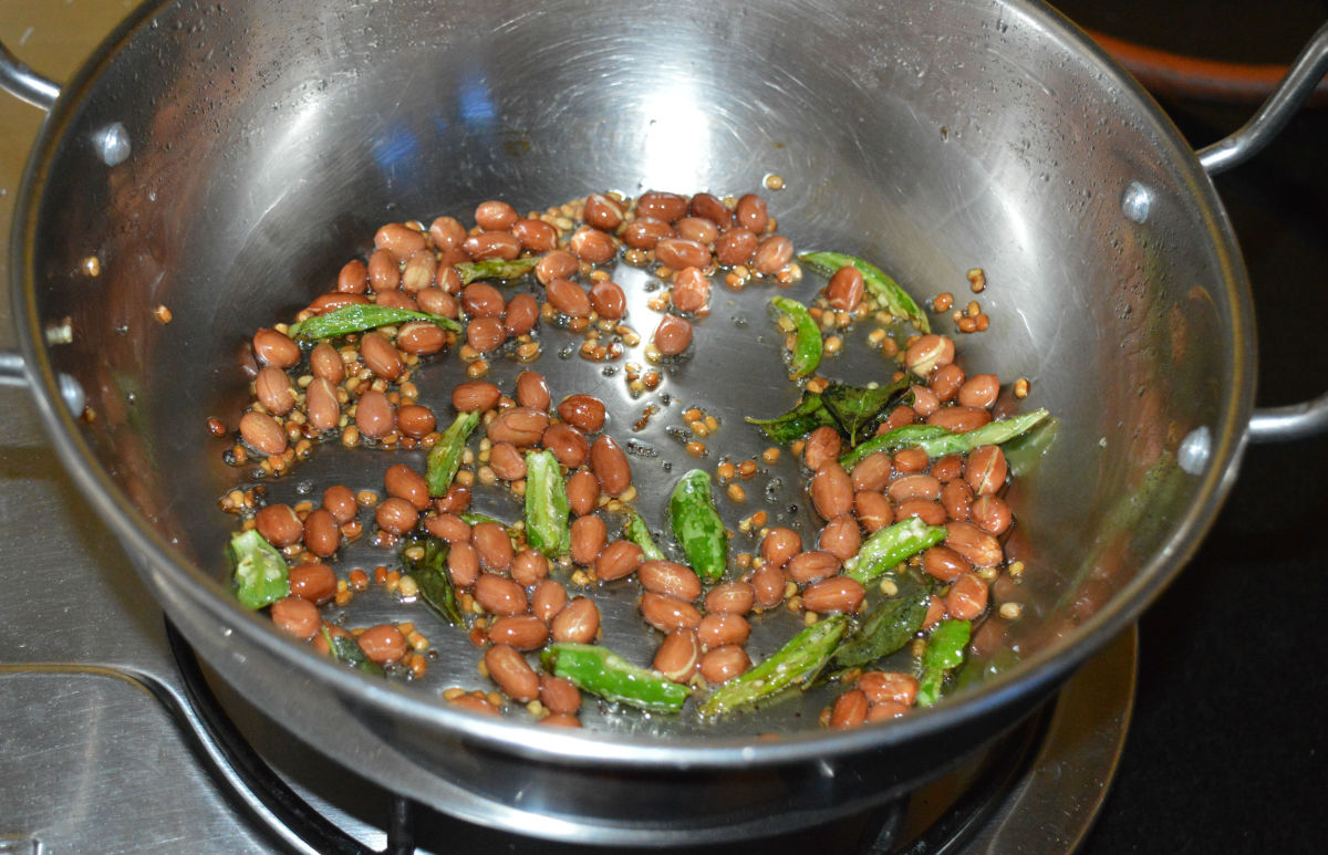 Step two: Heat oil in a deep-bottomed pan. Add mustard seeds. Let them crackle. Add chopped green chilies. Saute for a minute. Add peanuts, white lentils, and curry leaves. Saute until peanuts get crispy and lentils turn golden brown.