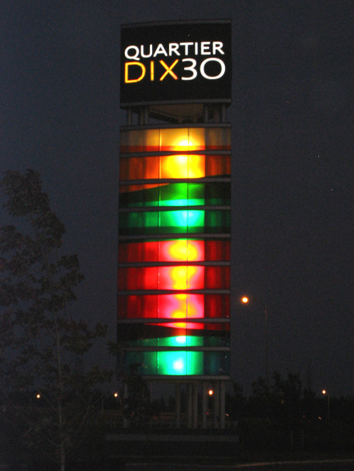 A photo of the illuminated Quartier DIX30 tower, seen at night; it is located at the eastern limits of the Quartier DIX30 development, adjacent to the Autoroute 10-30 cloverleaf. Brossard, Quebec, Canada.