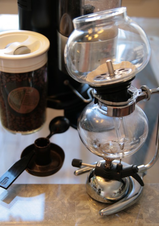 All the necessary components together: Hario siphon coffee maker, siphon paper filter, butane micro burner, coffee measuring spoon with stirrer, fresh roasted beans, Macap burr grinder.