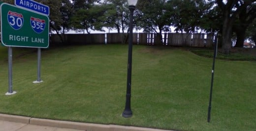 The grassy knoll from the street. A wood fence runs across the top of the small hill. Some witnesses thought shots came from this area. A polaroid photo taken from across the street showed a shadowy figure beneath the trees that wasn't there later.
