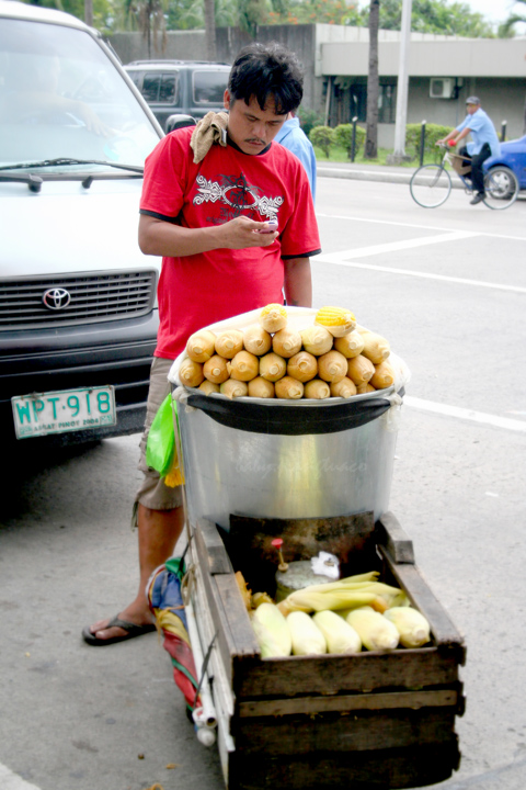 corn vendor texting somebody on his cell phone while waiting for buyers of his goods. Notice the boiled corn is kept hot by a portable stove underneath the big kettle