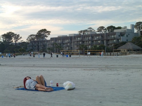 Lovers cuddle in the foreground as a group of young people play volleyball in the background.
