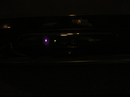 My Kinect device with lights off showing from left to right the red laser beam which can measure distance and height of users, the power indicator light, the camera and the IR light.  This camera is capable of taking pictures in the dark using IR.