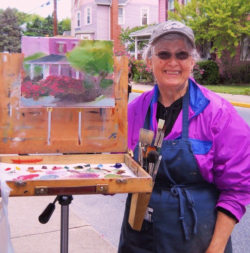 Mary Rinderle Smith standing next to her painting. The house she was painting is behind her and had colorful rose bushes in front of it. 