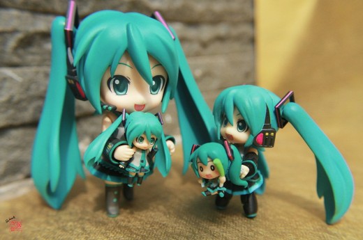 A "family" of Nendoroids. To the left we have a regular Nendoroid holding a Nendoroid Petit Plus. To the right we have a Nendoroid Petit holding a different Nendoroid Petit Plus. All of these figures are variations of the virtual singer Hatsune Miku.