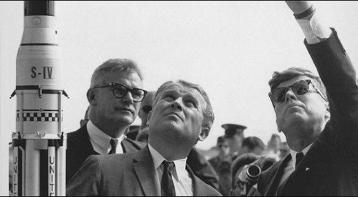 1961, actually a huge giant, JFK is on board with going to the moon and looking cool wearing shades. Here he plays the cool guy showing other giants which way the tiny human rocket will go to the moon. "Basically, up there, guys, real high up."