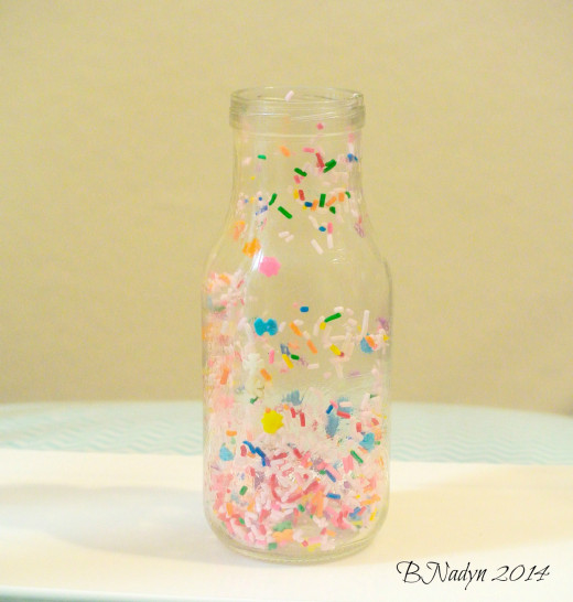 Start with coating the inside of the bottle with syrup or corn syrup so sprinkles will stick.  Pour in the sprinkles, rotating the bottle to spread them out.  Line rim with syrup and sprinkles, optional.