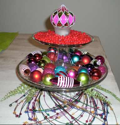 Two cake stands, stacked. Top cake stand has red hots surrounding an ornament upside down in a votive holder. Bottom plate has an assortment of ornaments.