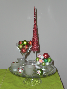 Glass cake plate with tree figurine, martini glass and small glass bowl filled with ornaments, votives and ornaments scattered.
