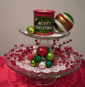 Two cake stands stacked. Top stand has 2 ornaments and a candle, bottom stand has wired berries and ornaments. Both stands are using doilies.