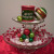 Two cake stands stacked. Top stand has 2 ornaments and a candle, bottom stand has wired berries and ornaments. Both stands are using doilies.
