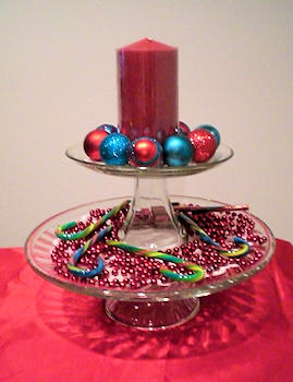 Stacked cake stands with 6" pillar candle surrounded by small ornaments on top and bead garland with candy canes on the bottom layer.