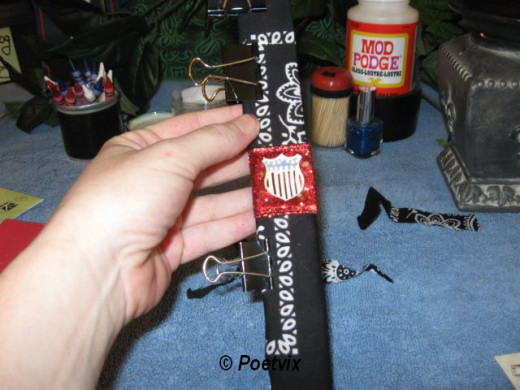 Bring in the ribbon and a coordinating sticker or two. Paint on your crayon. I used blue here on every third little paisley. When we take it through the laminating machine, it will remelt giving an interesting affect.