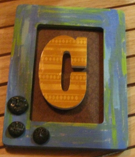 Give me a C! Framed art collage using a painted frame, scrap book paper covered letter, and three polymer clay faces.