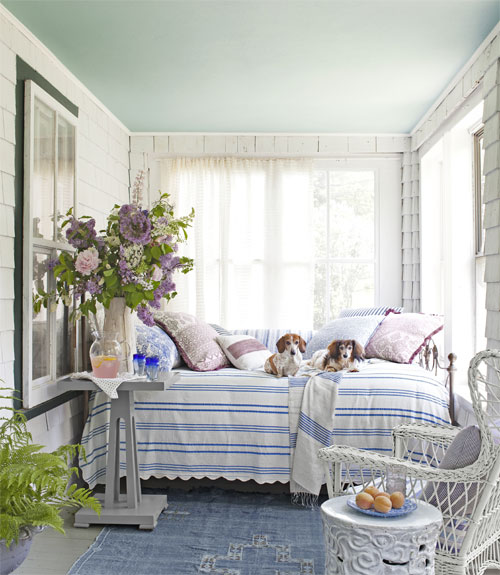Striped pastel blue &amp; white cover daybed nestled on the back porch. Oversized mauve flowers &amp; vase fit perfectly.  White furniture &amp; cloth blue rug.